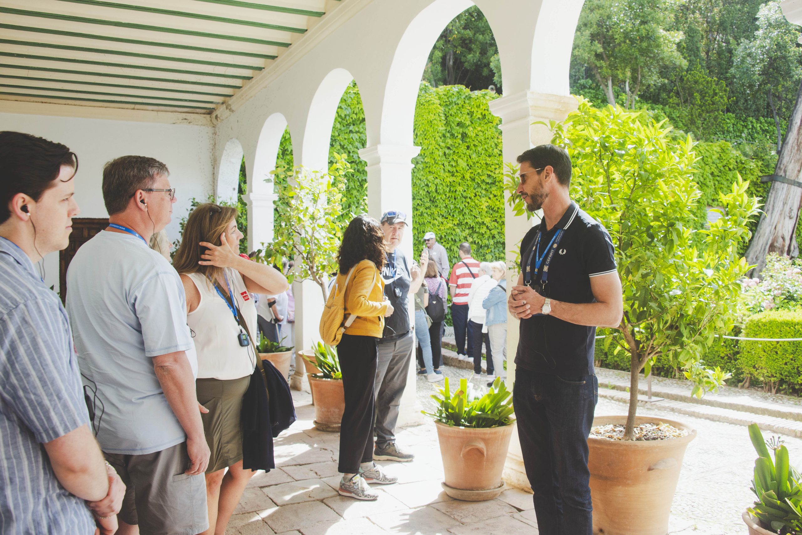 Un guía turístico conversa con un grupo de visitantes bajo una galería con arcos blancos de medio punto, decorada con macetas y plantas en un entorno monumental.