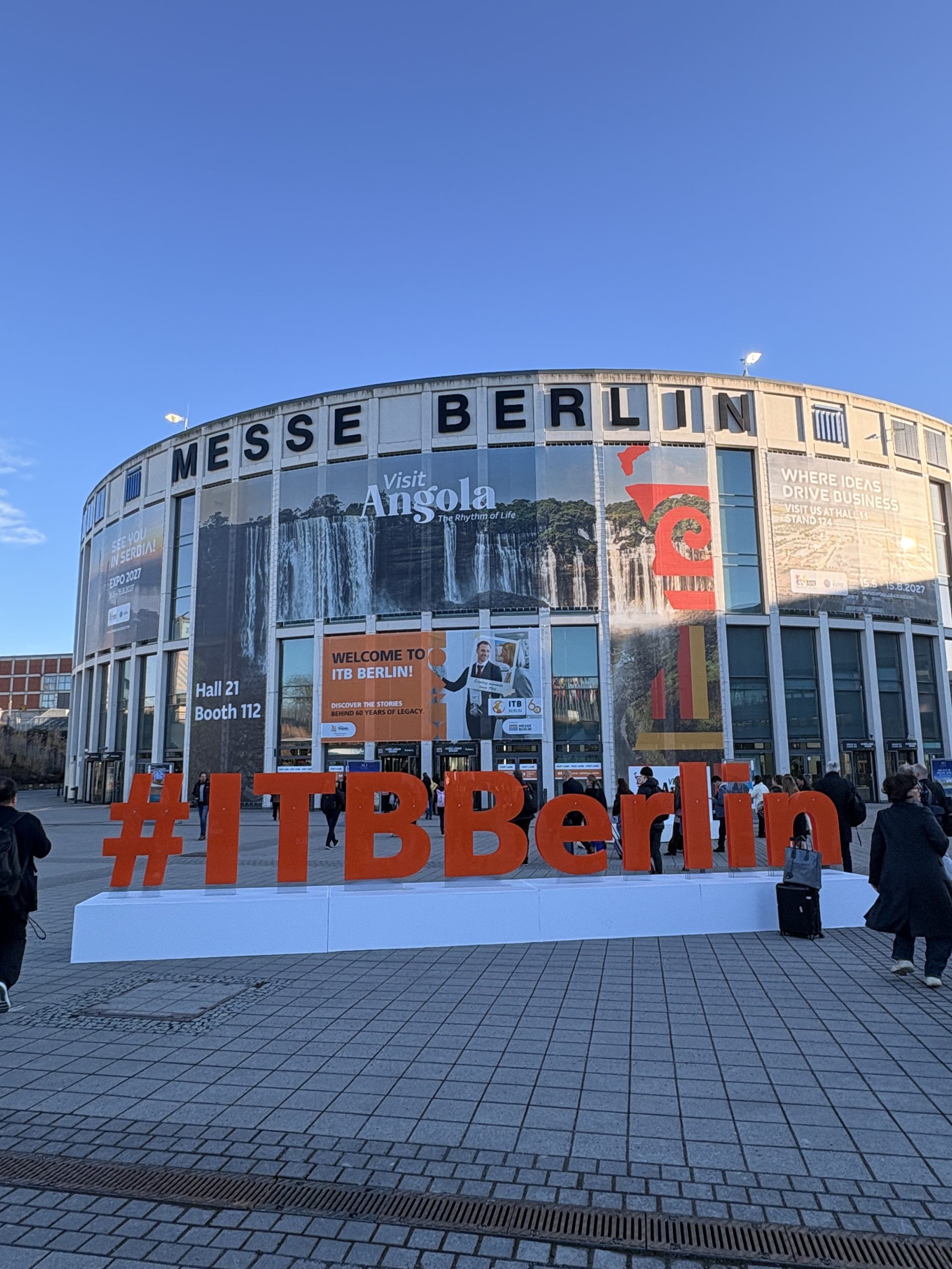 Exterior architectural view of the iconic Messe Berlin circular building with the official ITB Berlin travel trade show hashtag promotion displayed in the foreground.