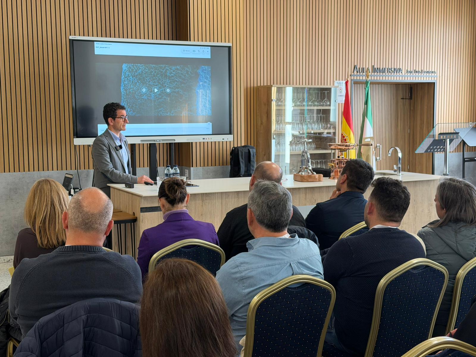 Asistentes siguiendo una presentación con imagen ampliada en una sala formativa con banderas oficiales.
