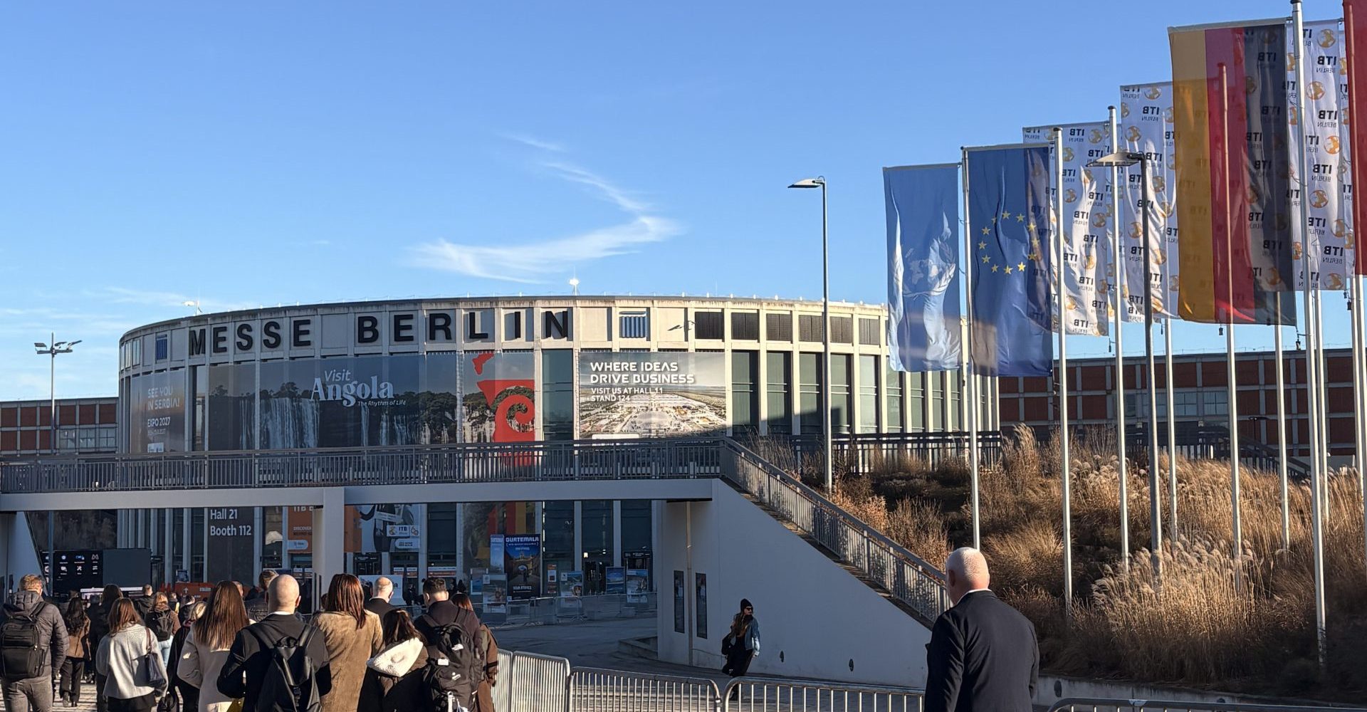 Exterior view of the iconic Messe Berlin exhibition center entrance with international flags flying on a clear sunny day during a major global travel industry fair.