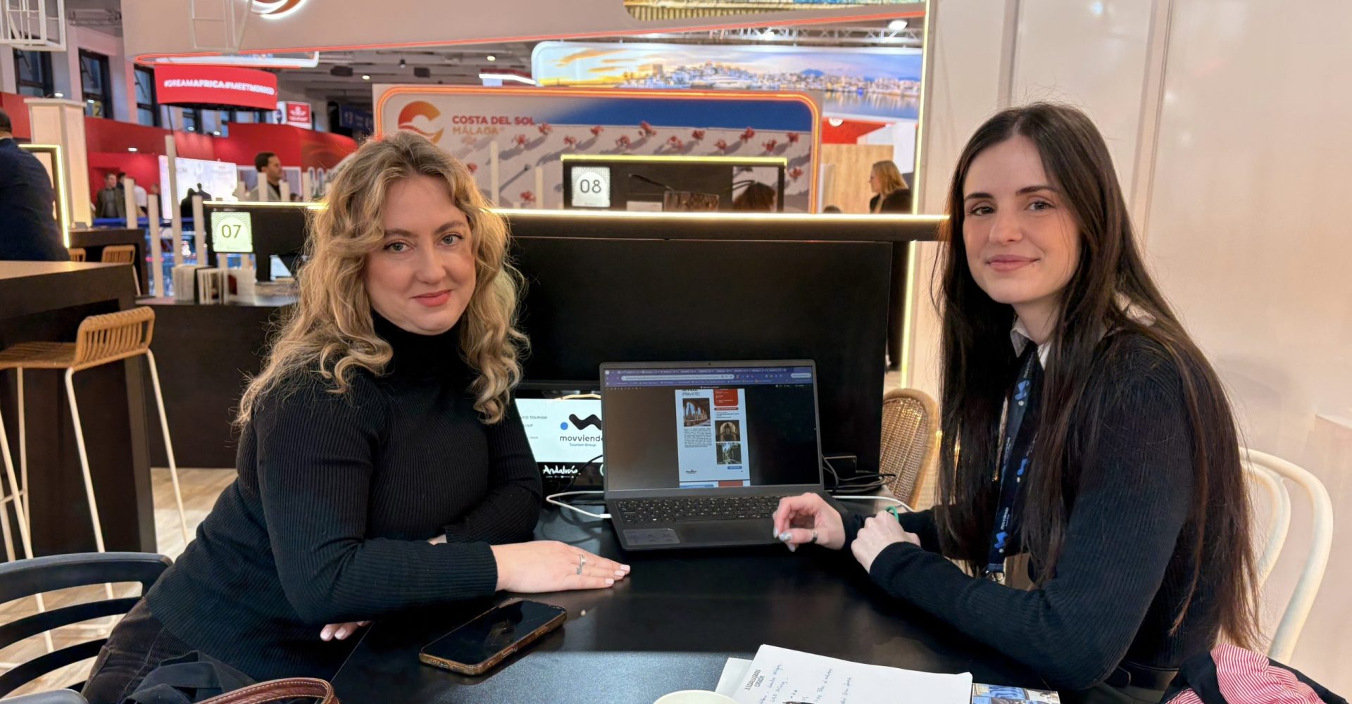 Two professional female corporate representatives discussing regional travel projects and DMC logistics at the Costa del Sol promotional desk during a business forum.
