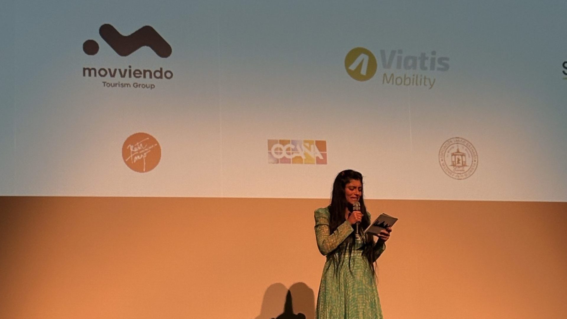 A female corporate speaker giving an institutional presentation on a stage with official Andalusian tourism brand logos projected on the background screen.