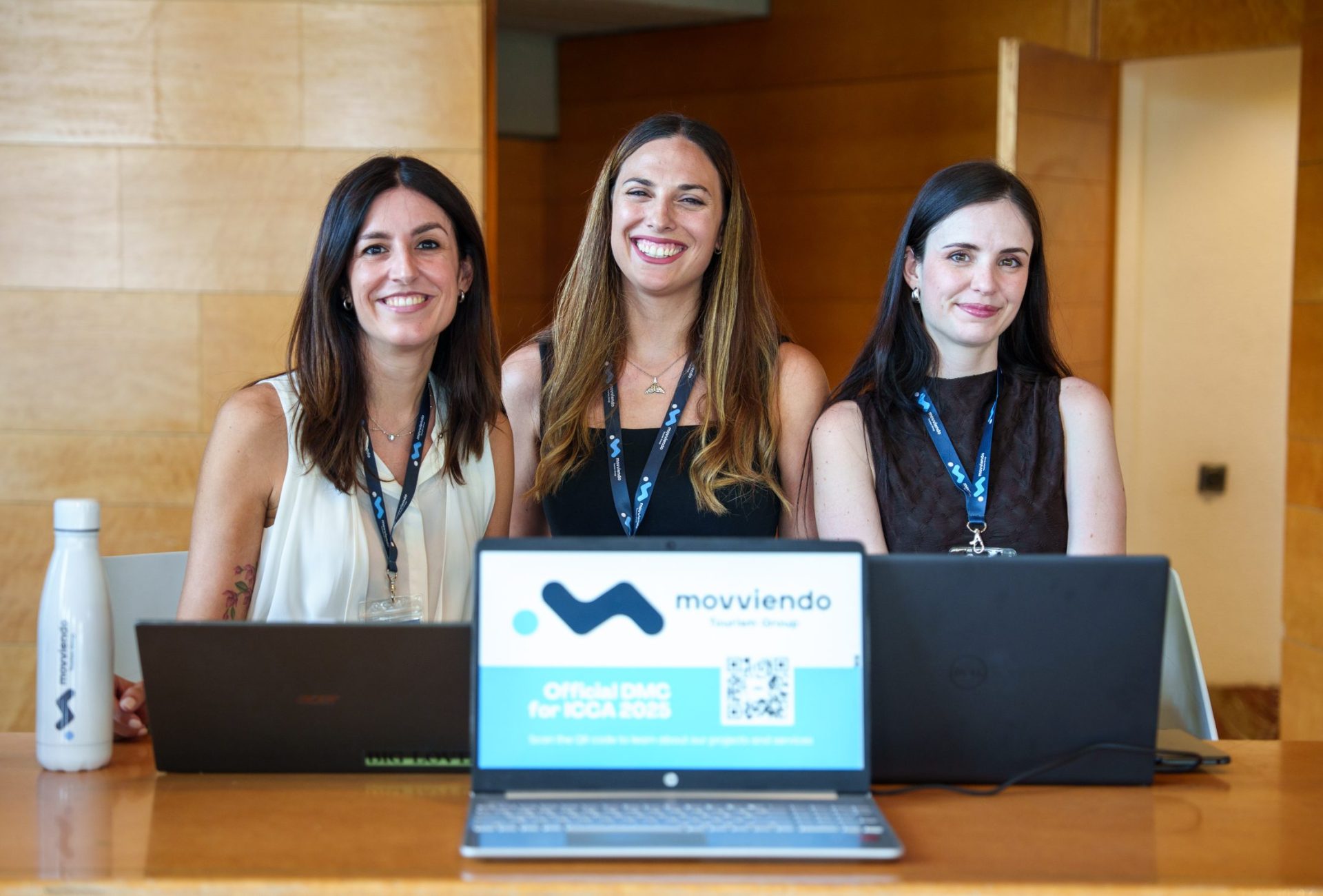 Three smiling professional business managers posing together during a corporate meeting with an open laptop showing the company brand logo.