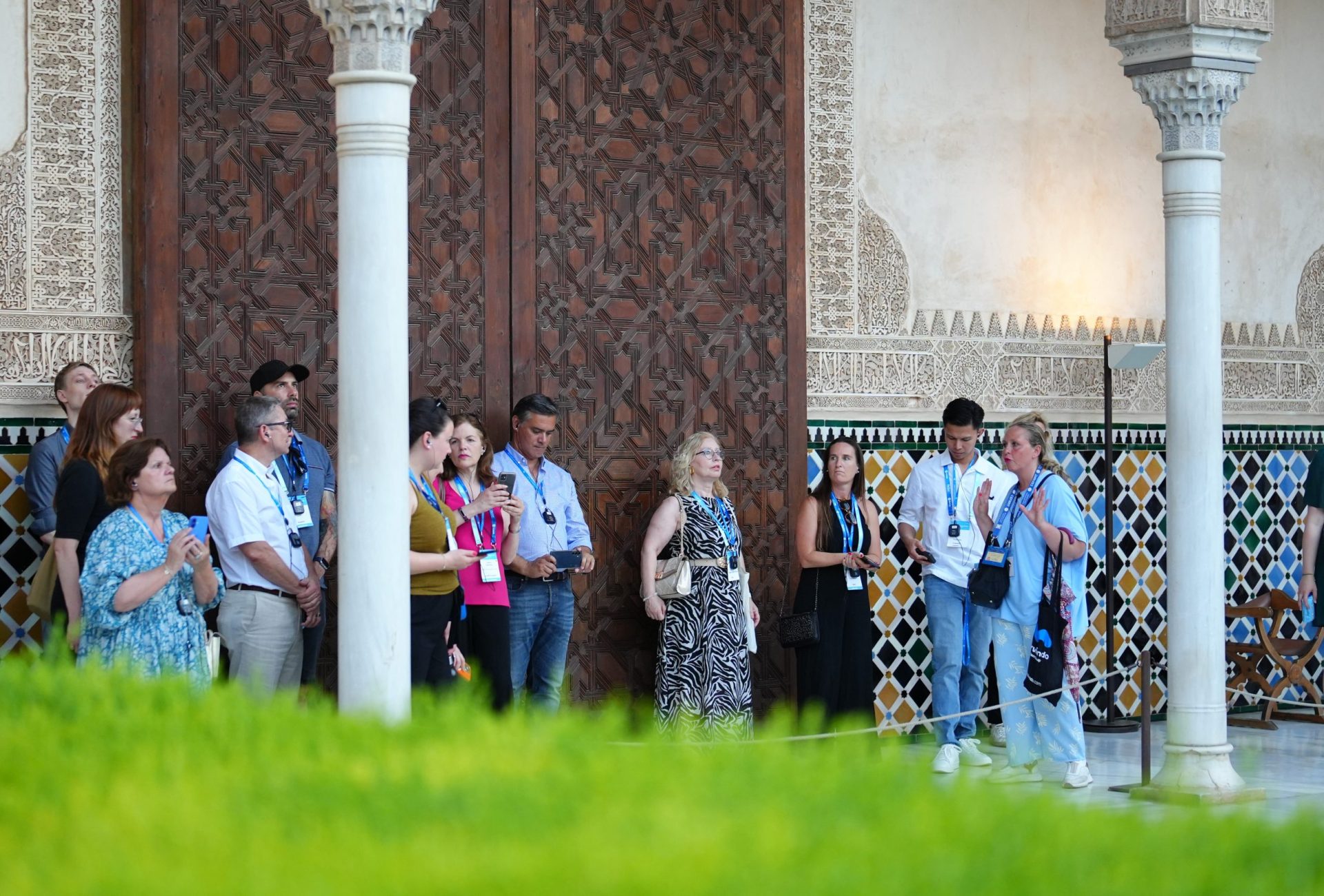 un grupo de personas realiza una visita guiada en los exteriores de los Palacios Nazaríes de la Alhambra de Granada, con detalles arquitectónicos de fondo.