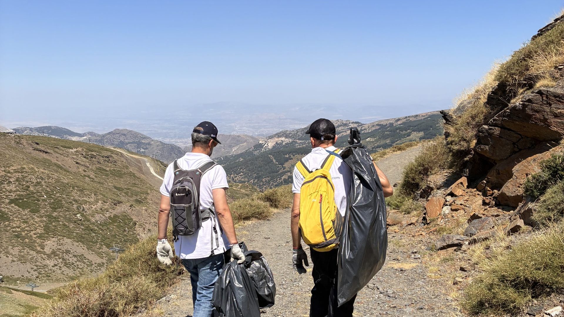 Three young environmental volunteers with backpacks walking along a scenic high-mountain trail during a corporate social responsibility CSR sustainability day.