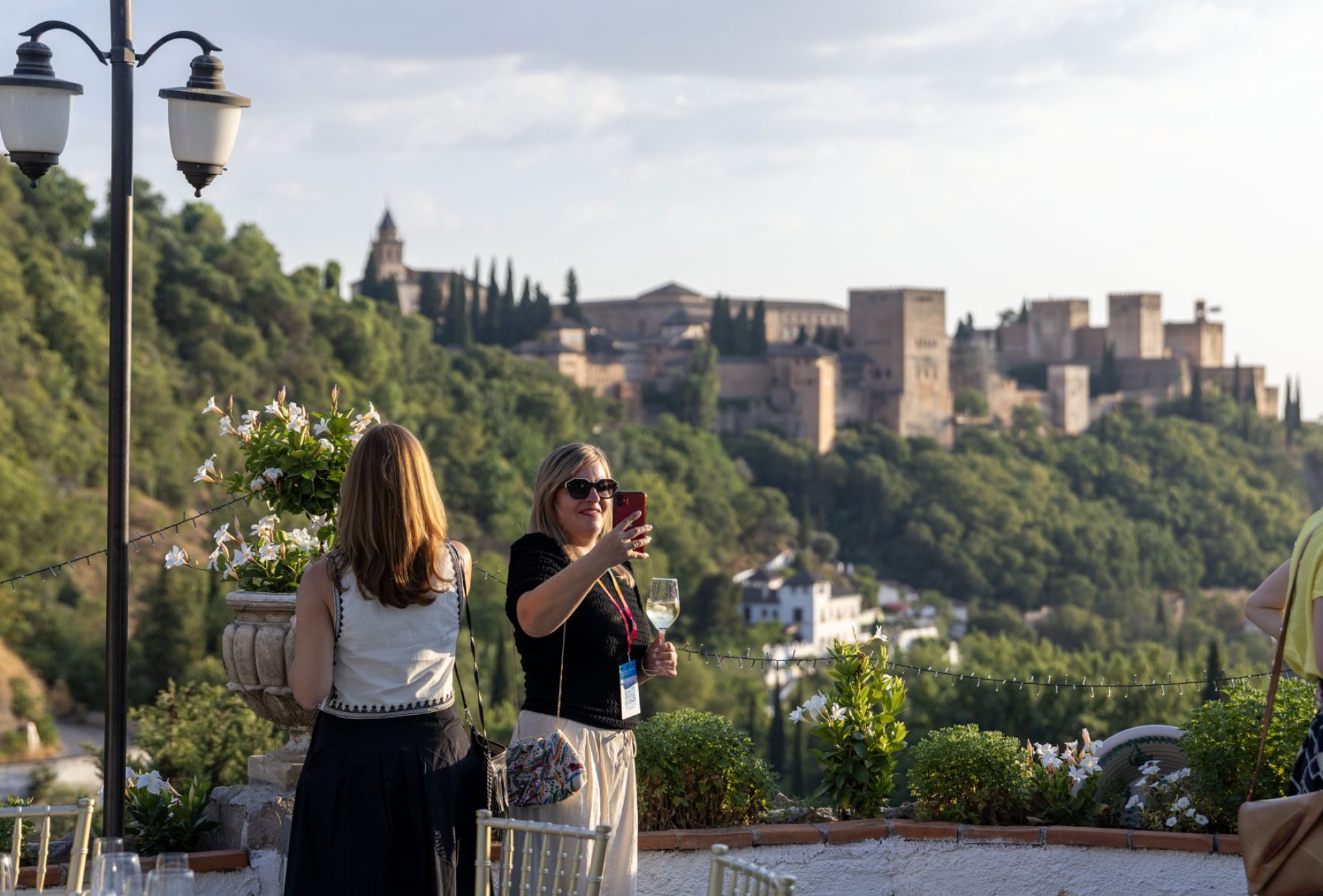 Un grupo de turistas contempla y hace fotografías de las vistas de la ciudad de Granada y las montañas al fondo desde un mirador empedrado durante el atardecer.