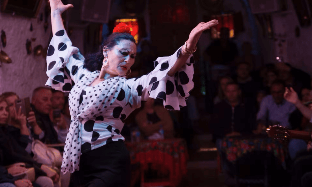 Bailaora de flamenco con vestido blanco de lunares negros realiza una pose con los brazos elevados en un tablao iluminado.