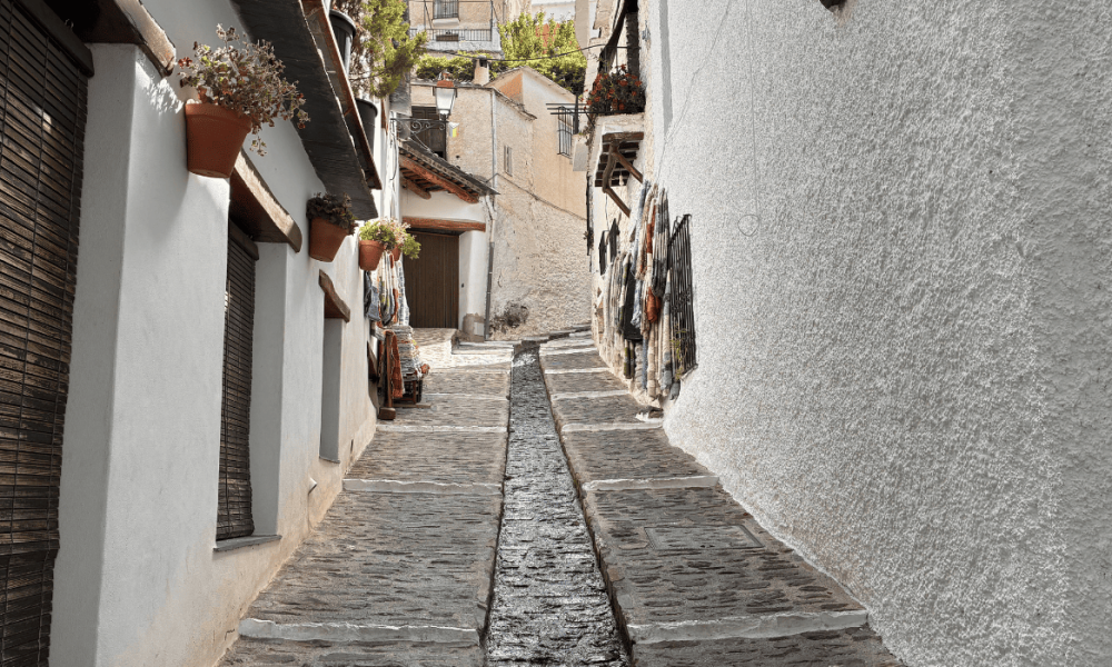 Calle peatonal estrecha de piedra con una persona caminando entre paredes blancas y rejas de ventanas.