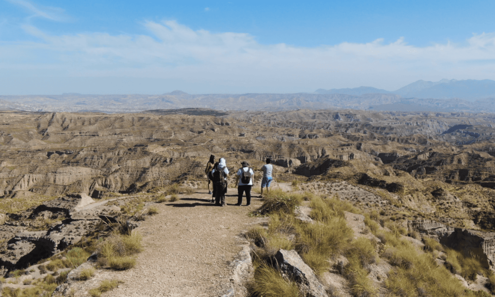 Personas caminan por un sendero de tierra rodeado de formaciones geológicas áridas de color marrón en el Geoparque de Granada.