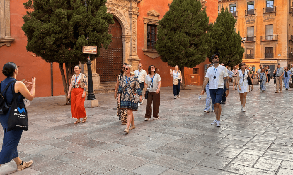 Personas caminando por una plaza de piedra frente a la fachada de un edificio histórico con árboles al fondo.