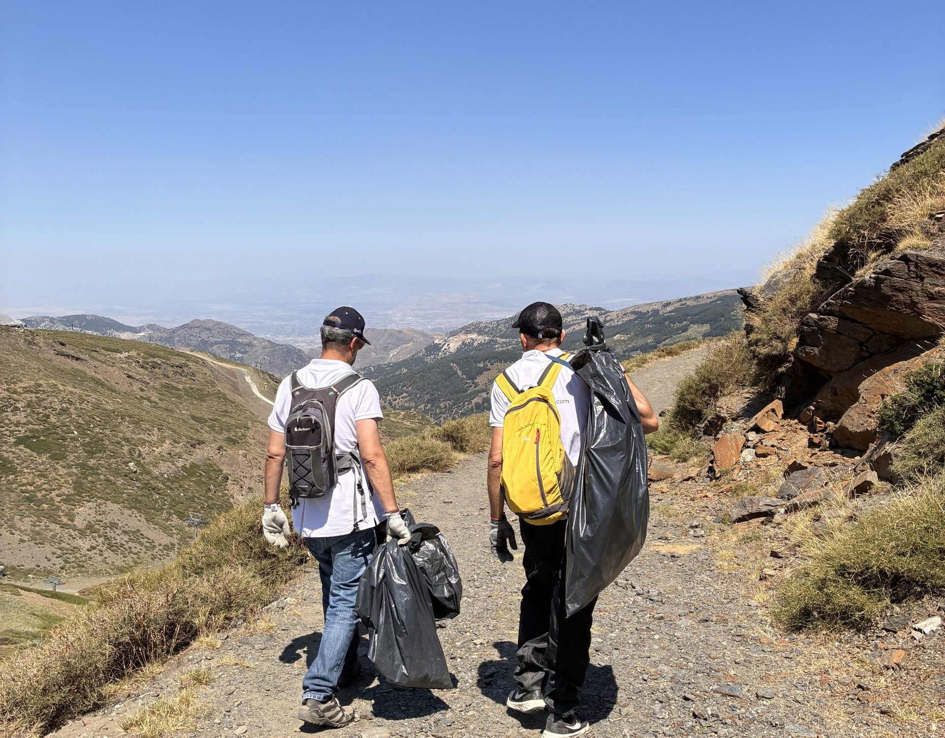 Tres jóvenes voluntarios con mochilas recorriendo un sendero de alta montaña bajo un cielo despejado en una actividad de sostenibilidad.